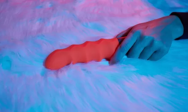 Uma Mulher Deitada Em Uma Cama, Segurando Um Vibrador Em Forma De Dildo Na Mão, Parecendo Relaxada E Engajada Em Autoexploração.