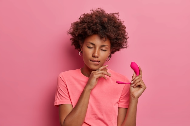 Uma Mulher De Pele Escura, Satisfeita, Morde Os Lábios E Fecha Os Olhos De Prazer Enquanto Segura Um Vibrador Inteligente De Silicone Com Controle Por Aplicativo, Contra Uma Parede Rosa.