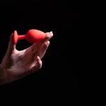 Um Par De Mãos Femininas Segurando Um Plug Anal Vermelho Contra Um Fundo Preto, Com Espaço Amplo Para Texto.