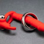 Close-up De Um Vibrador Anal Vermelho E Um Anel Peniano De Metal Sobre Um Fundo Cinza, Ilustrando Opções De Brinquedos Sexuais.