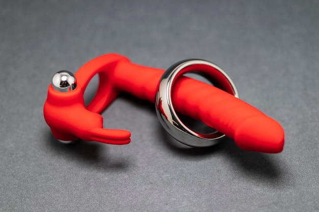 Close-up De Um Vibrador Anal Vermelho E Um Anel Peniano De Metal Sobre Um Fundo Cinza, Ilustrando Opções De Brinquedos Sexuais.