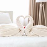 Uma Colcha Macia Adornada Com Dois Cisnes De Toalha E Pétalas De Rosa Espalhadas Em Um Quarto De Hotel Bem Iluminado.
