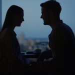 Um Jovem Casal Se Beijando E Se Abraçando Em Um Ambiente De Escritório, Com Janelas Panorâmicas Ao Fundo.