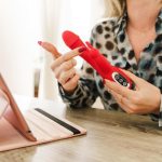 Uma Jovem Blogueira Gravando Um Vlog Enquanto Revisa Um Brinquedo Sexual, Sentada Em Uma Mesa Com Um Fundo Colorido.