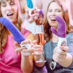 Duas Mulheres Animadas Em Uma Loja De Sex Shop Vibrante, Cada Uma Segurando Um Dildo E Uma Bebida, Cercadas Por Decorações Coloridas.