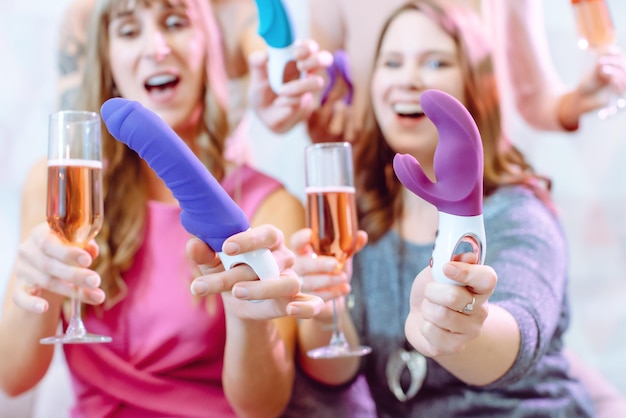 Duas Mulheres Animadas Em Uma Loja De Sex Shop Vibrante, Cada Uma Segurando Um Dildo E Uma Bebida, Cercadas Por Decorações Coloridas.